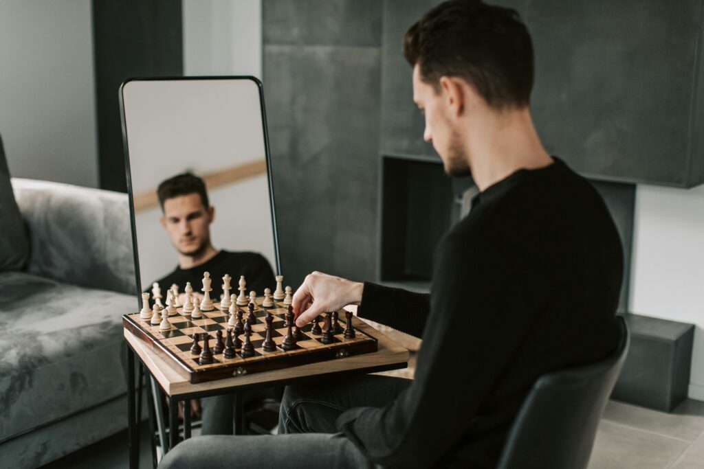 A man plays chess on a wooden board with a reflection in a mirror indoors.