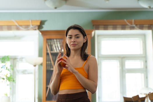 A young woman in a tank top holds an apple inside a bright, well-lit room with a bookcase backdrop.